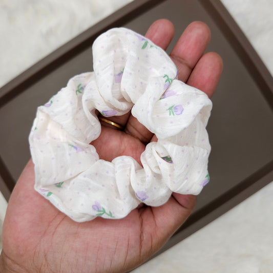 Hand holding a white scrunchie with floral patterns on a neutral background