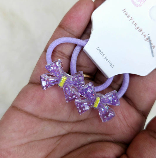 Purple hair tie with glittery bows held by a hand, with a brand label in the background.