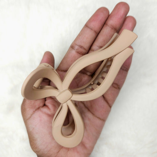 Beige bow-shaped matte hair claw clip held in a hand against a white background