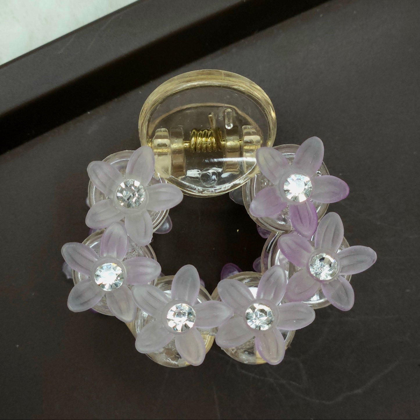 A petite, round lavender hair claw that showcases an overlay of opaque and translucent purple flower beads and crystal accents.