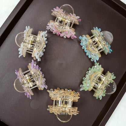 A top-down shot shows six small, circular hair claw clips arranged on a dark brown tray. Each clip is adorned with translucent, flower-shaped beads and tiny sparkling rhinestones, and they come in various pastel colors: white, pink, light blue, lavender, peach, and light green.