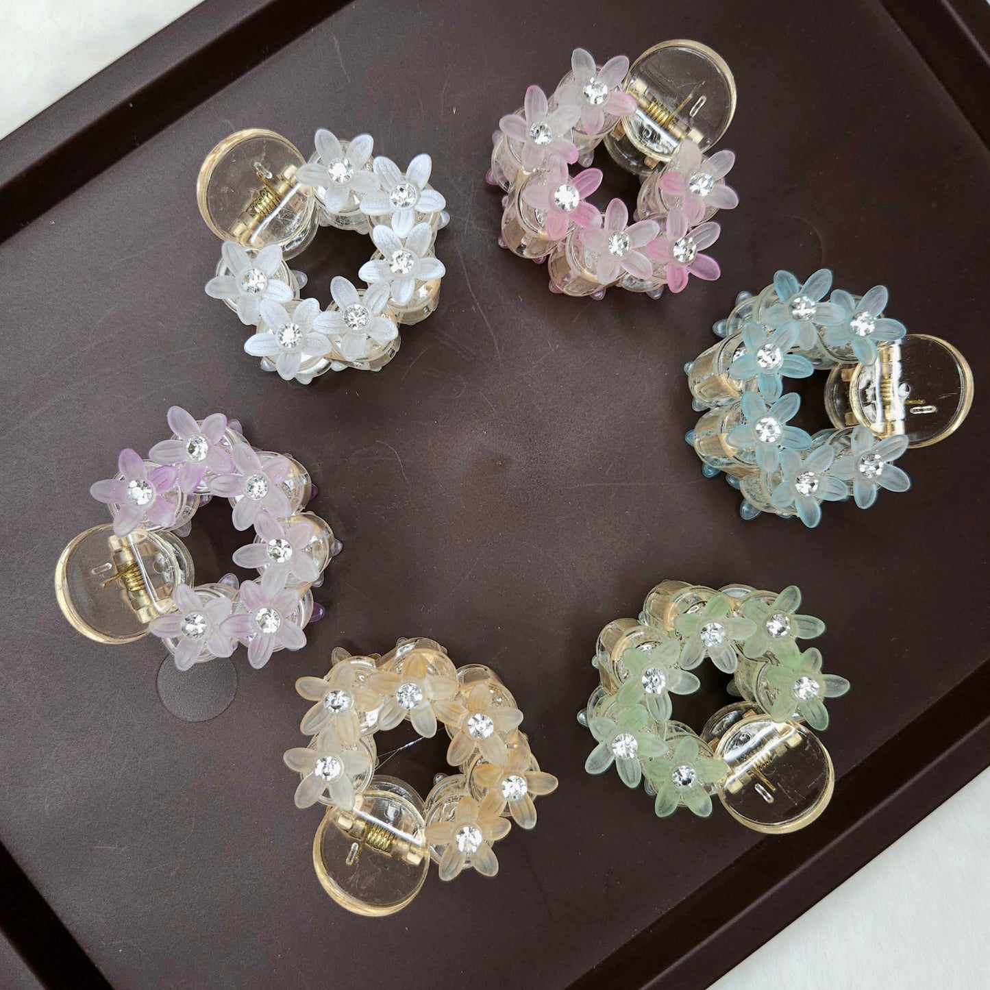 A top-down shot shows six small, circular hair claw clips arranged on a dark brown tray. Each clip is adorned with translucent, flower-shaped beads and tiny sparkling rhinestones, and they come in various pastel colors: white, pink, light blue, lavender, peach, and light green.
