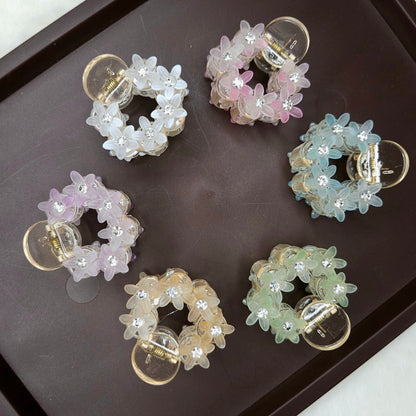 A top-down shot shows six small, circular hair claw clips arranged on a dark brown tray. Each clip is adorned with translucent, flower-shaped beads and tiny sparkling rhinestones, and they come in various pastel colors: white, pink, light blue, lavender, peach, and light green.