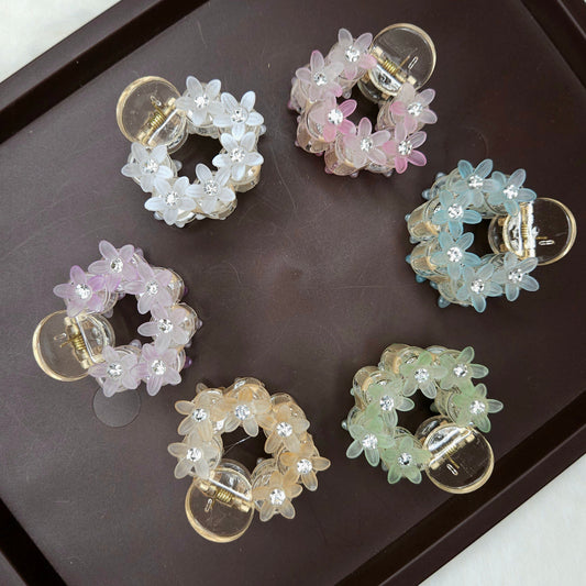 A top-down shot shows six small, circular hair claw clips arranged on a dark brown tray. Each clip is adorned with translucent, flower-shaped beads and tiny sparkling rhinestones, and they come in various pastel colors: white, pink, light blue, lavender, peach, and light green.