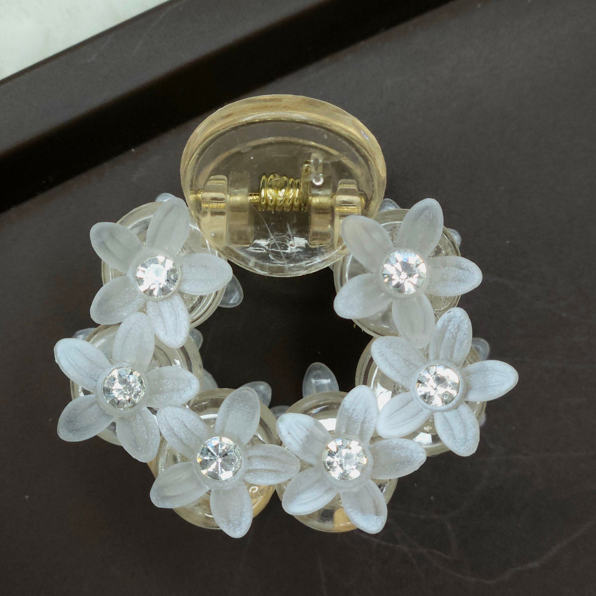 Close-up of a tiny, circular white flower hair claw clip adorned with translucent petals and a scattering of delicate rhinestones.