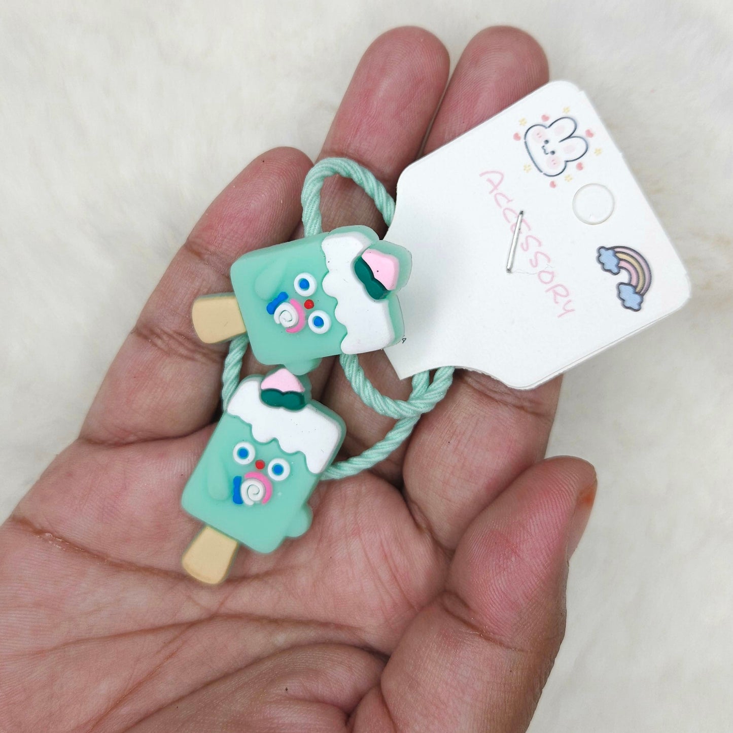 Hand holding a small green ice cream-shaped hair tie with a tag on a light background