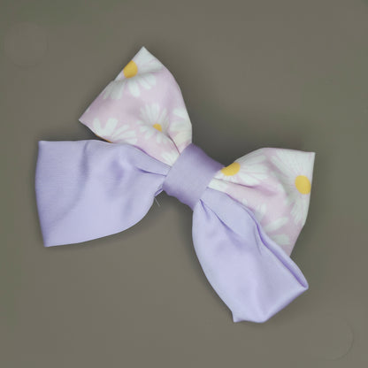 A close-up of a lavender satin hair bow featuring a white daisy floral print, with a slight sheen on the fabric.