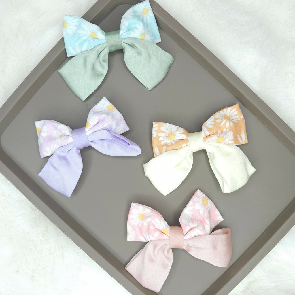 Multiple daisy-printed satin hair bows, including a pink, a blue, and a cream one, laid out to show their glossy texture and classic bow shape.