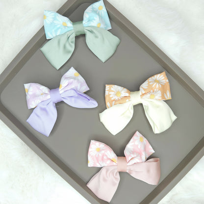 Multiple daisy-printed satin hair bows, including a pink, a blue, and a cream one, laid out to show their glossy texture and classic bow shape.