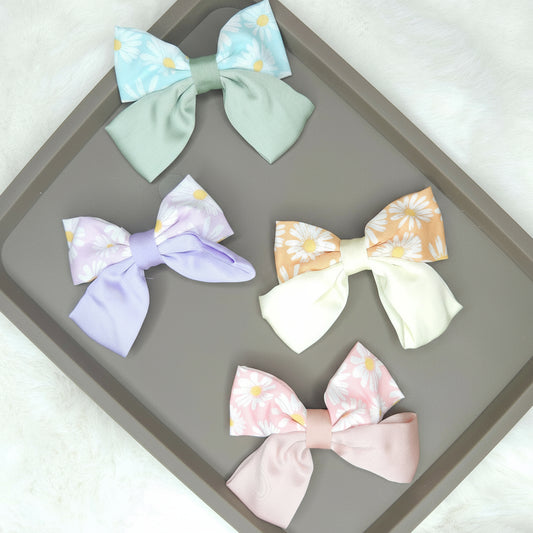 Multiple daisy-printed satin hair bows, including a pink, a blue, and a cream one, laid out to show their glossy texture and classic bow shape.