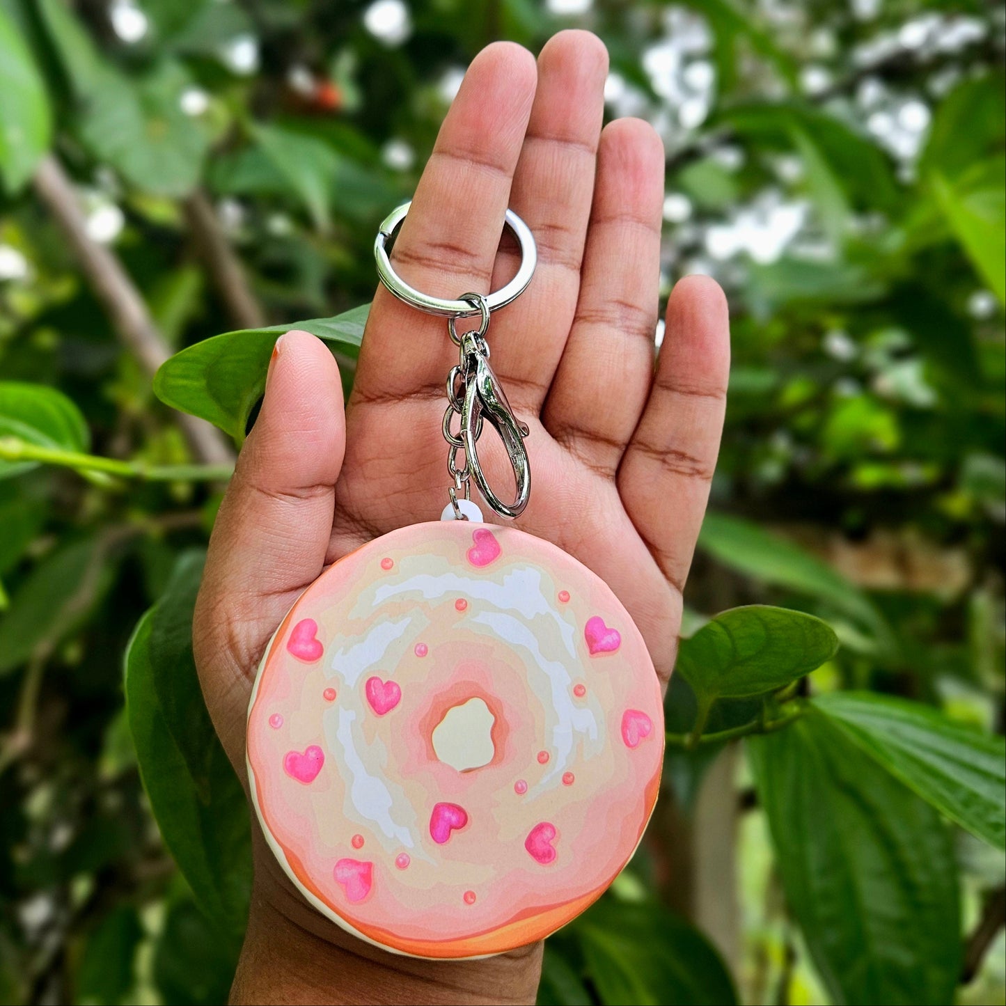 Hand holding a keychain shaped like a donut with pink icing and sprinkles against a green leafy background.