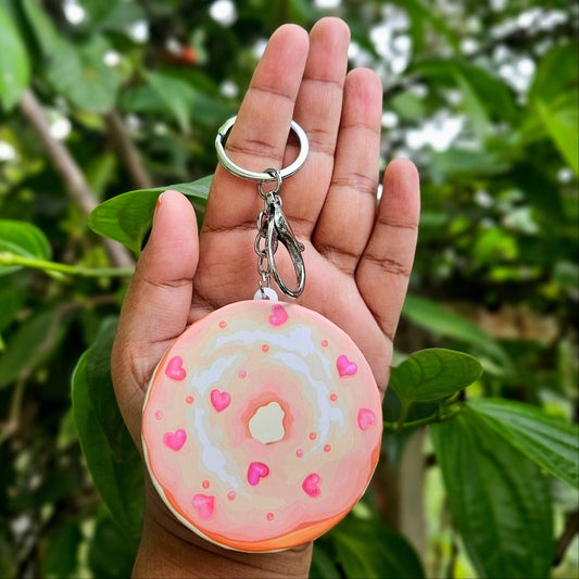 Hand holding a keychain shaped like a donut with pink icing and sprinkles against a green leafy background.