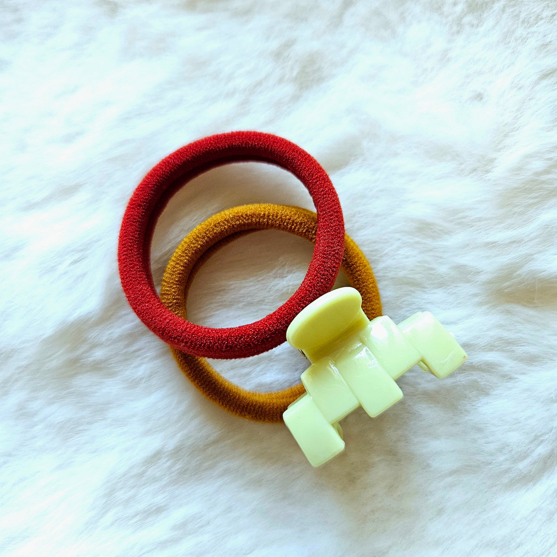 Two hair ties, one red and one yellow, with a white toy figure on a white fur background.