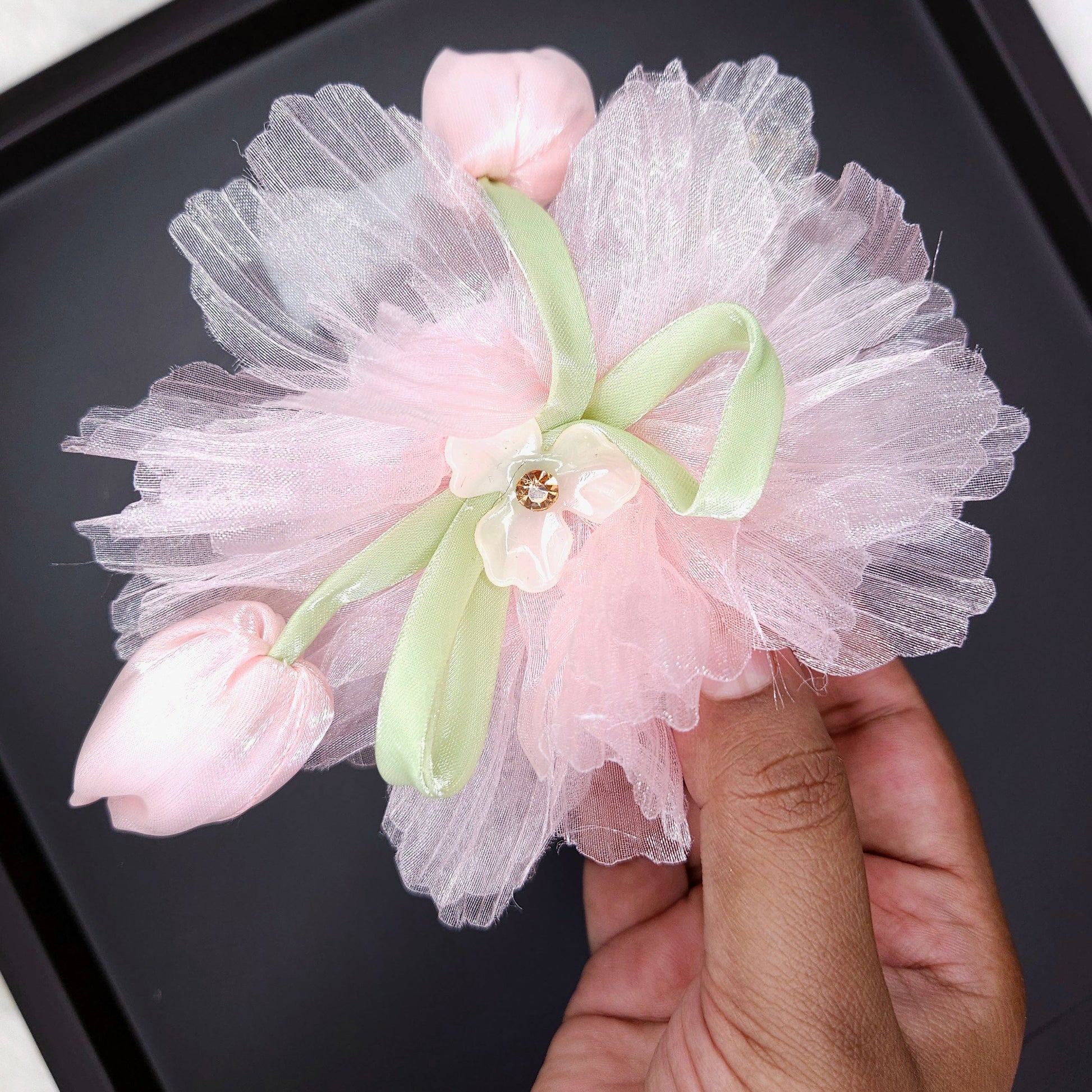 Decorative flower accessory with tulle and pink buds held by a hand on a dark surface.