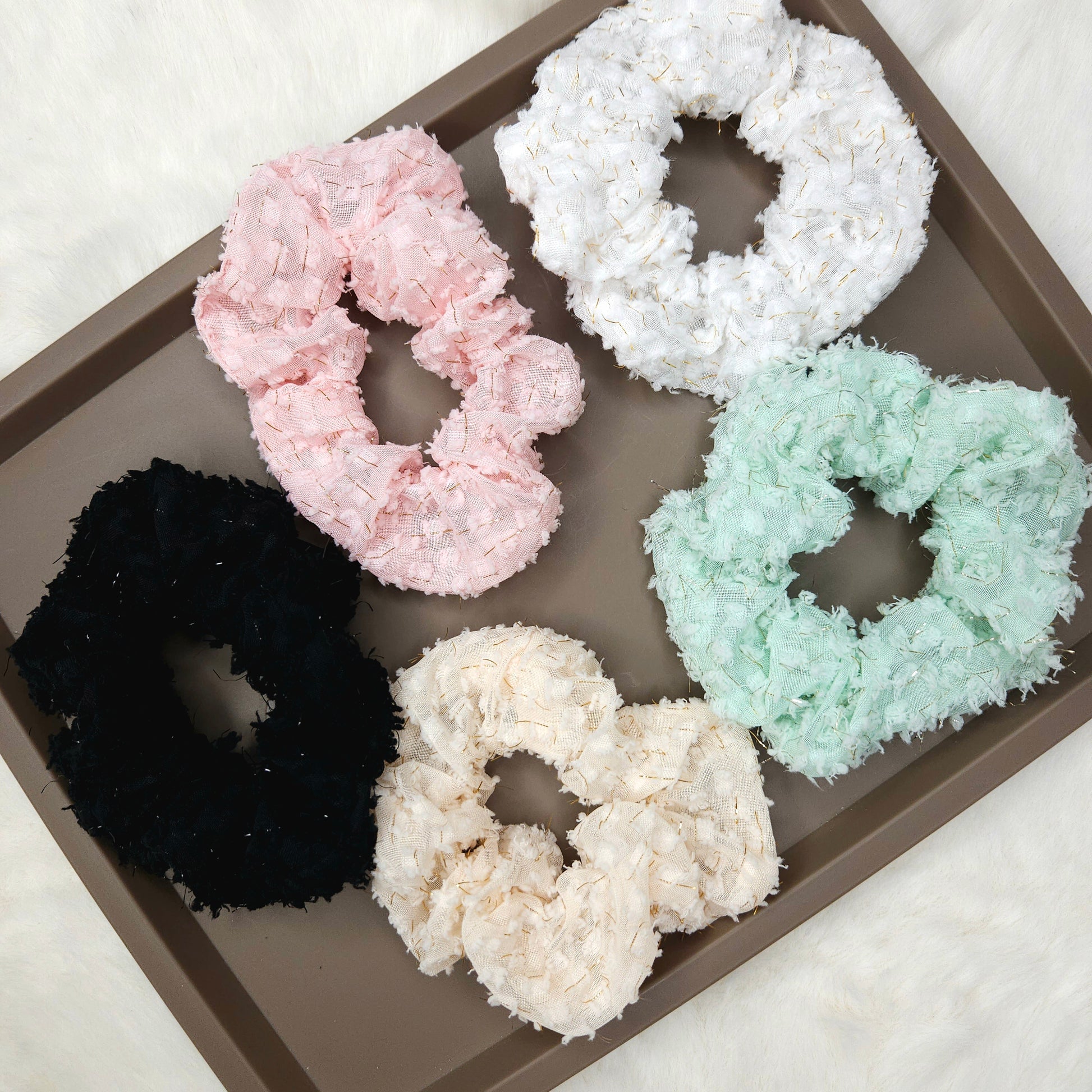 Five floral textured scrunchies in pink, white, black, and green on a brown tray.
