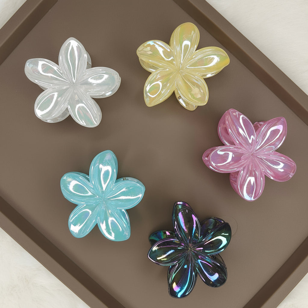 A set of medium-sized flower-shaped hair claw clips in a variety of glimmering colors, such as iridescent pink, gold, and silver, laid out together on a surface.
