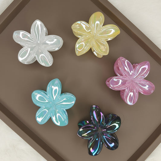 A set of medium-sized flower-shaped hair claw clips in a variety of glimmering colors, such as iridescent pink, gold, and silver, laid out together on a surface.