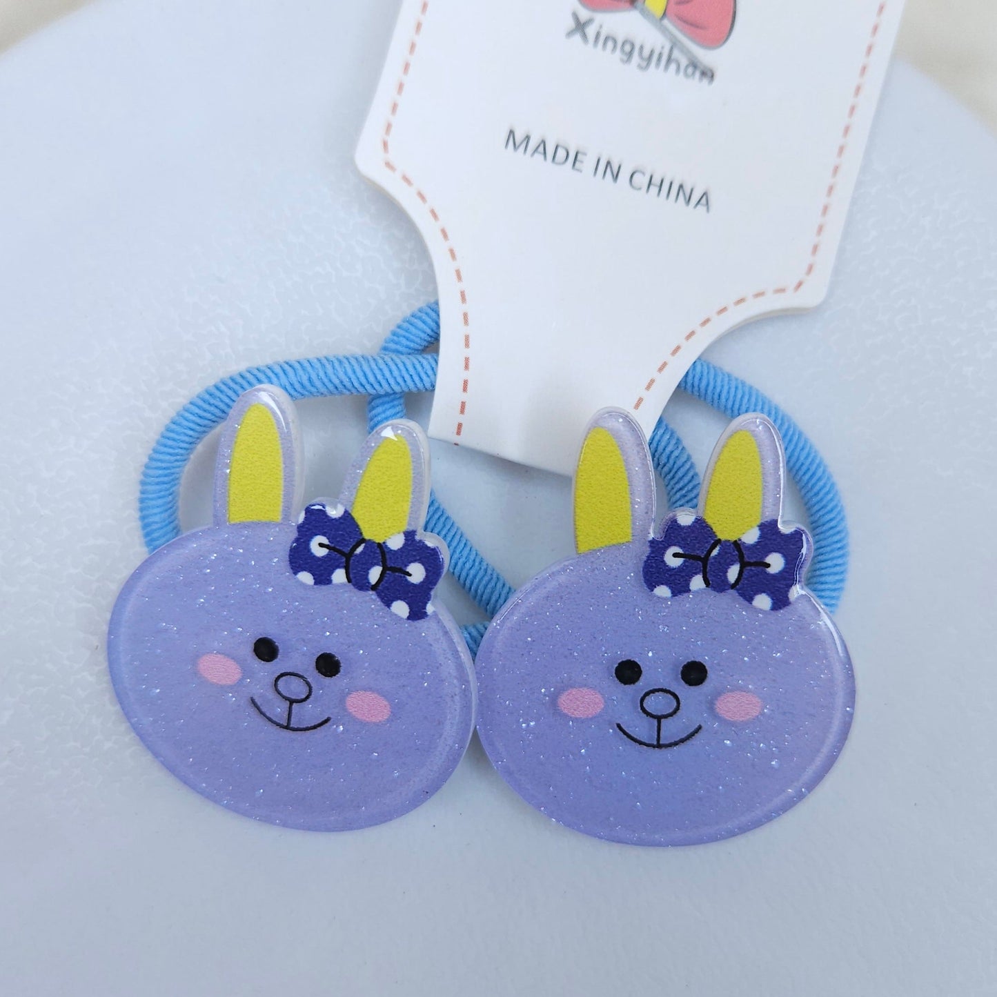 Two purple hair ties bunny ears with blue bows on a white background, branded 'Xingyihua'.