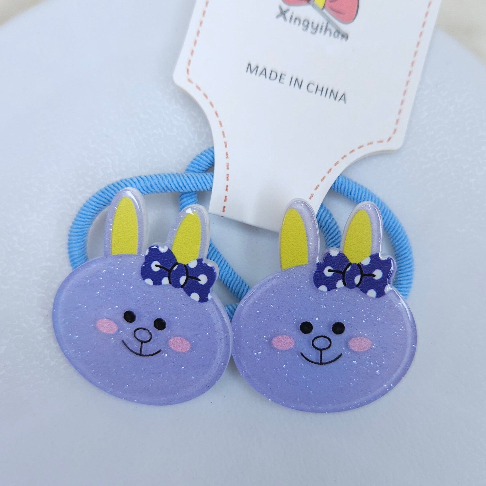Two purple hair ties bunny ears with blue bows on a white background, branded 'Xingyihua'.