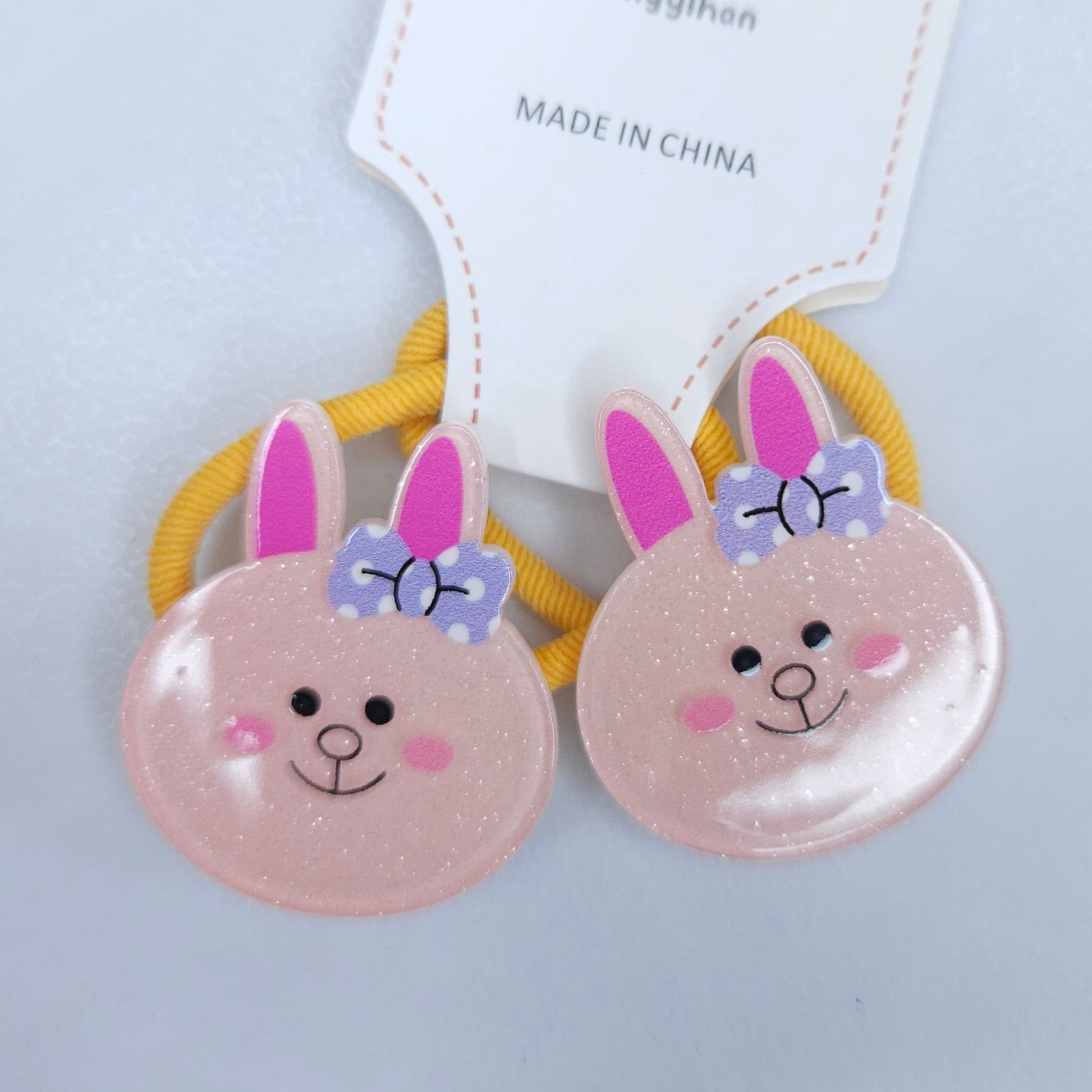 Two pink bunny face hair ties with yellow bands on a light gray background, branded 'Xingyihan'.
