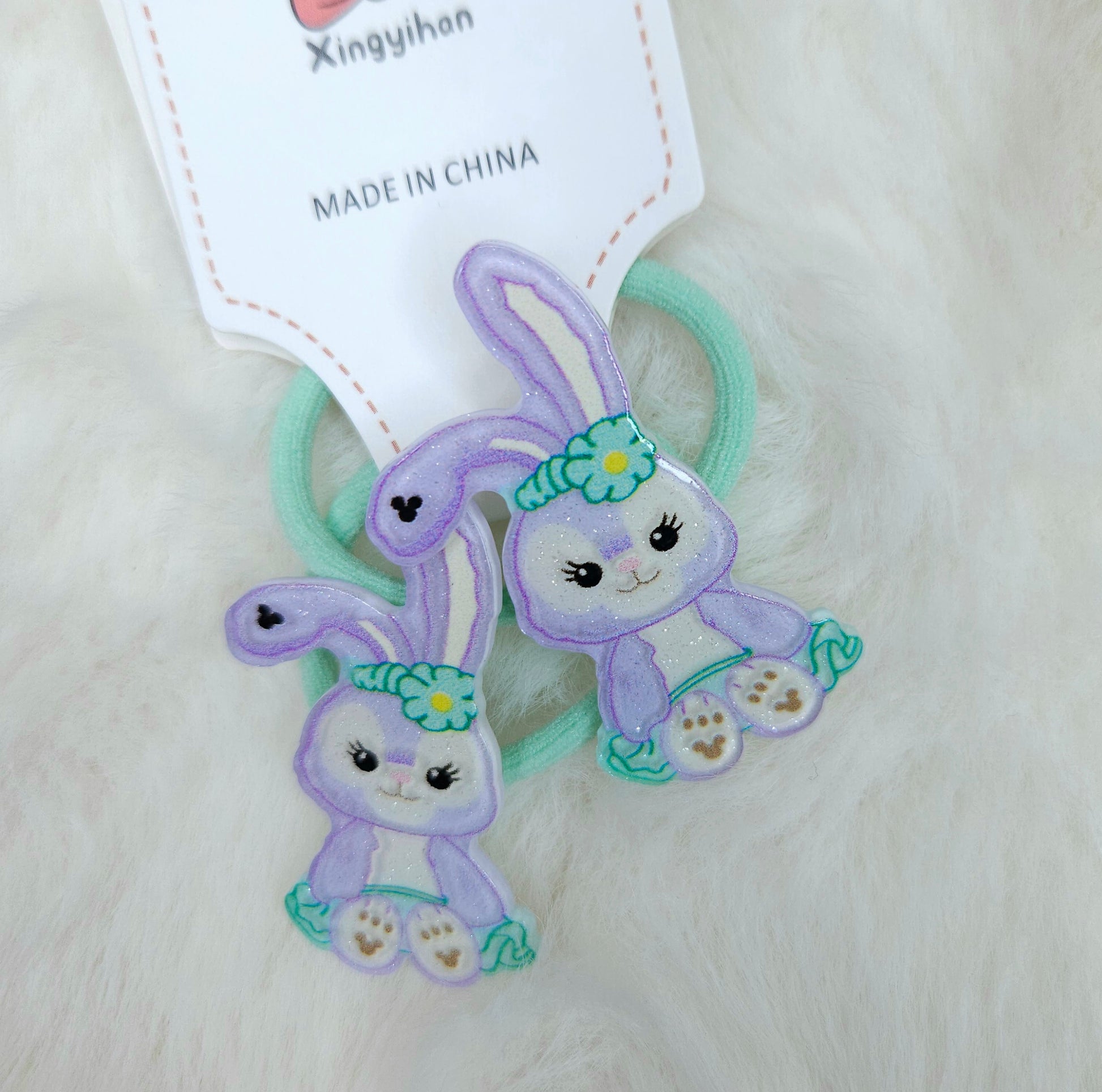 Two purple animal-shaped hair ties with green ribbons on a white background, accompanied by a 'Made in China' label.