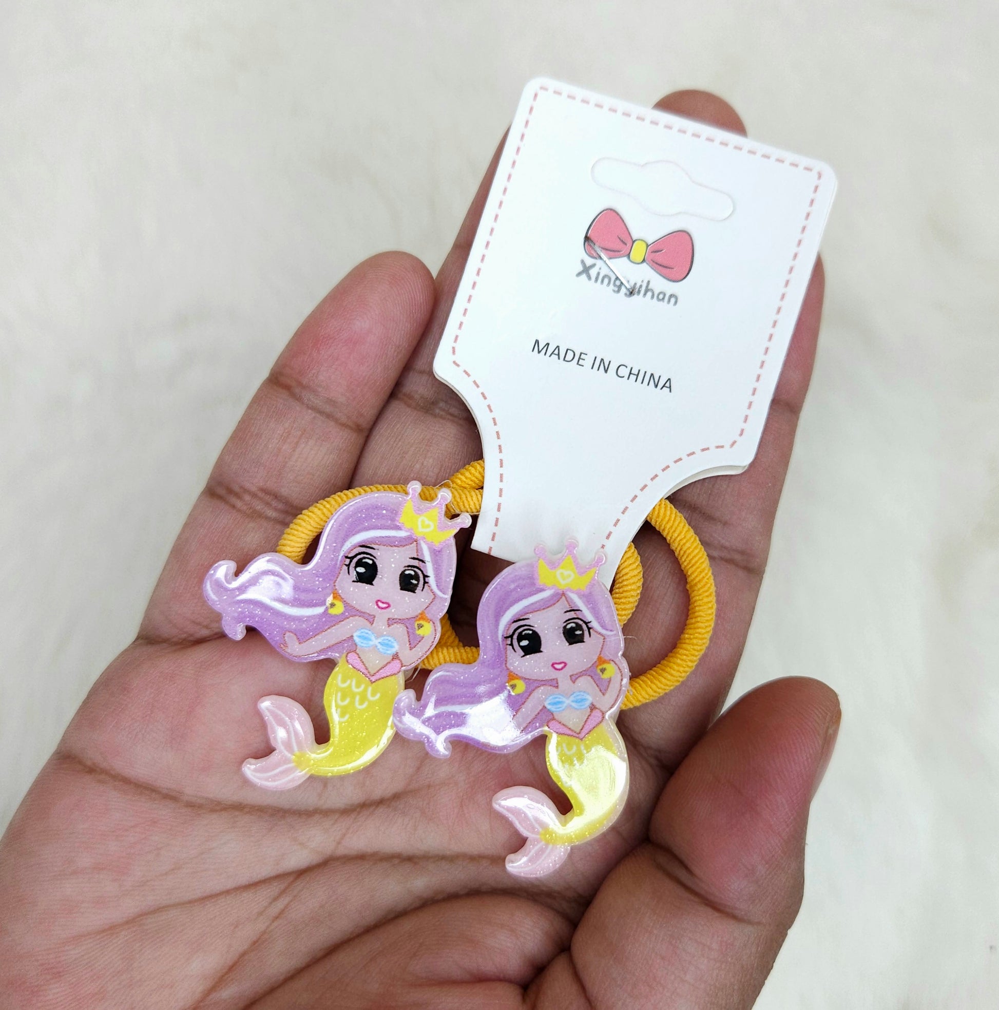 Two mermaid-shaped hair ties held by a hand with a 'Xingyihuan' branded card.