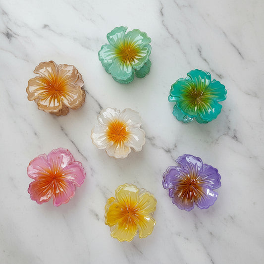 Overhead photo of seven iridescent, transparent floral hair claw clips for women, with colors like pastel pink, lavender, amber, and seafoam green, arranged on a white fluffy background.