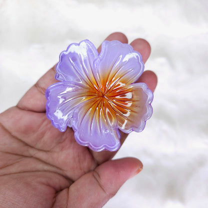 Close-up of a hand holding a large purple and white translucent floral hair claw clip with an orange-yellow center, against a fluffy white background.