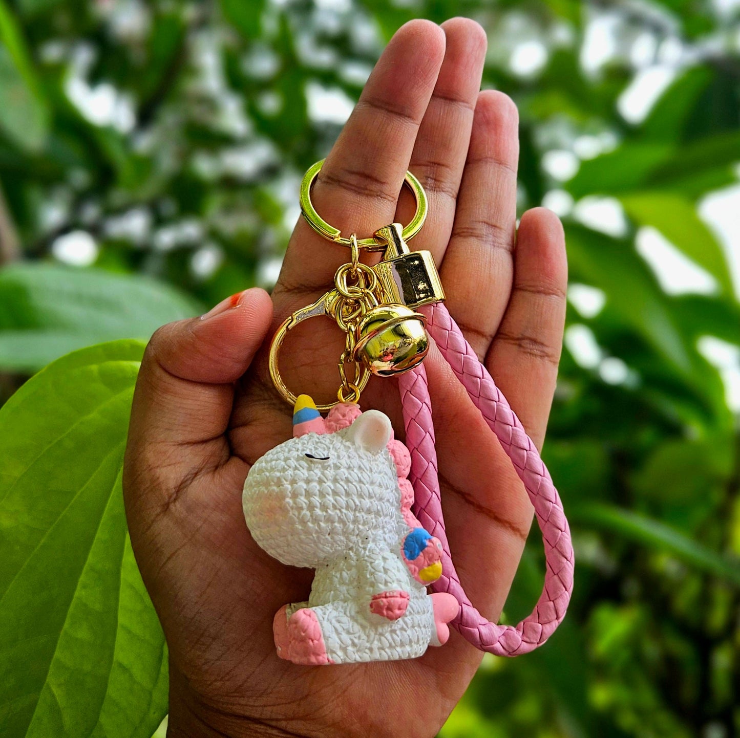 Hand holding a keychain with a small plush toy attached, against a green leafy background