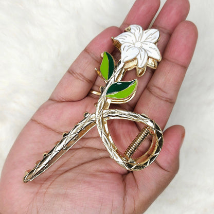 Gold hair clip with a white flower and green leaves held in a hand against a white background