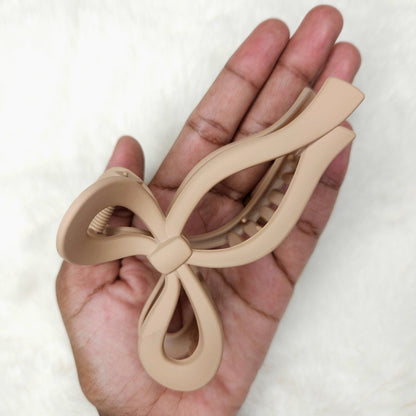 Beige bow-shaped matte hair claw clip held in a hand against a white background