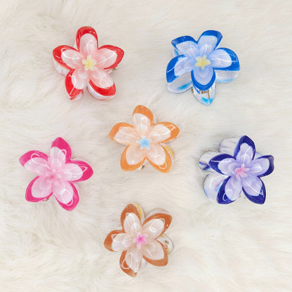 Six colorful translucent plastic flower-shaped hair clips are arranged on a white fluffy surface. From left to right, top row: red and white, blue and white. Middle row: pink and white, orange and white, purple and white. Bottom row: brown and white.