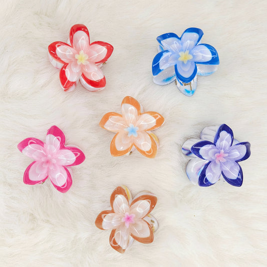 Six colorful translucent plastic flower-shaped hair clips are arranged on a white fluffy surface. From left to right, top row: red and white, blue and white. Middle row: pink and white, orange and white, purple and white. Bottom row: brown and white.