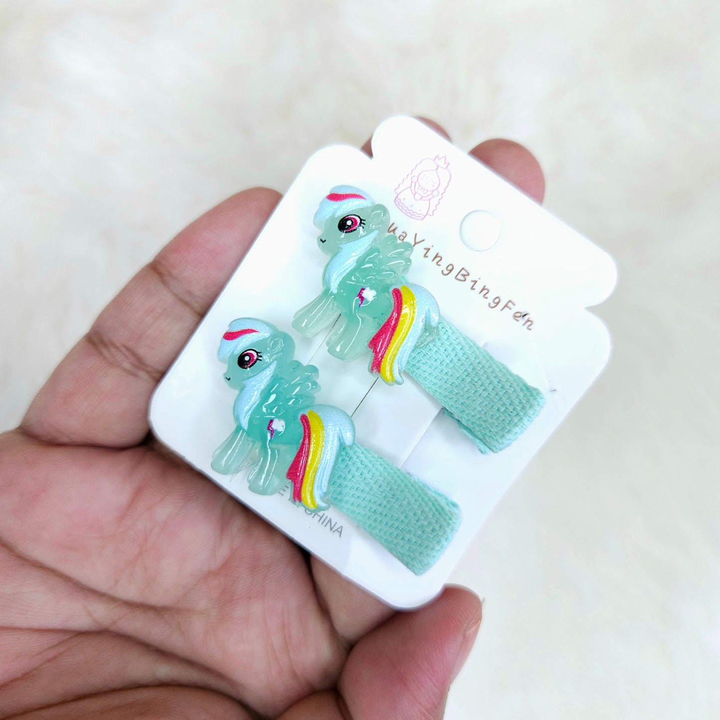Hand holding a set of green hair clips with rainbow designs on a white card.
