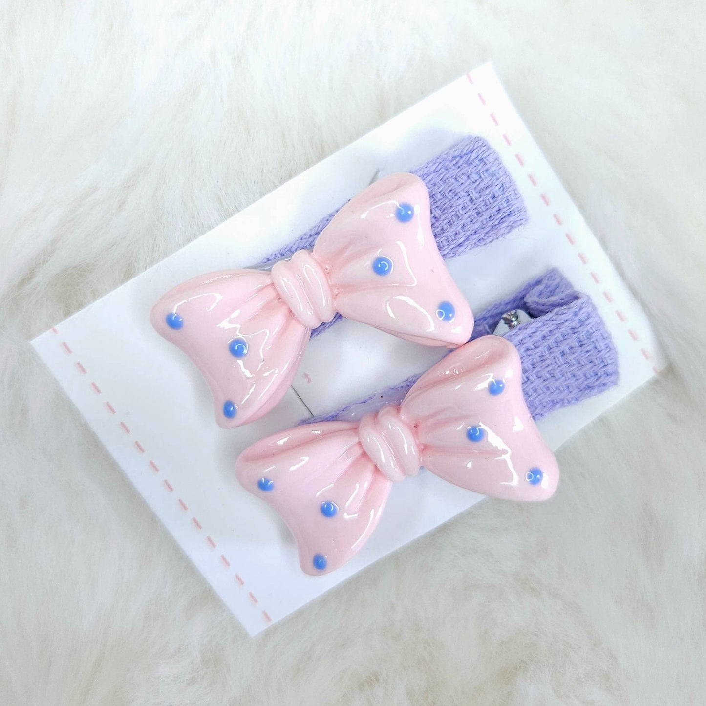 Two pink and purple hair bows with polka dots on a white card against a light gray background.