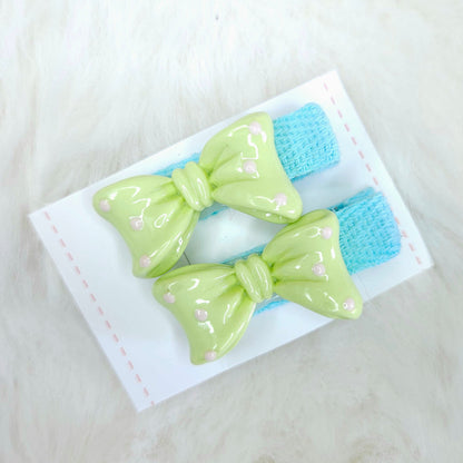 Two green hair bows with blue centers on a white card against a white background