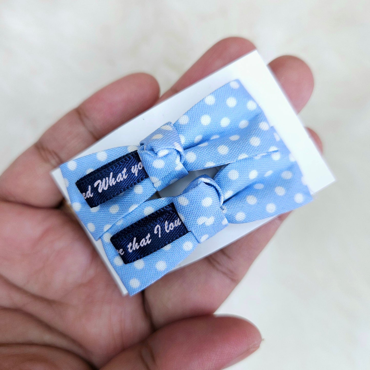 Two blue polka dot hair bows with text on a white box held in a hand.