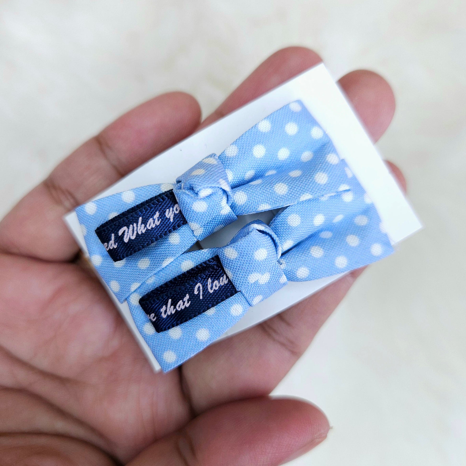 Two blue polka dot hair bows with text on a white box held in a hand.