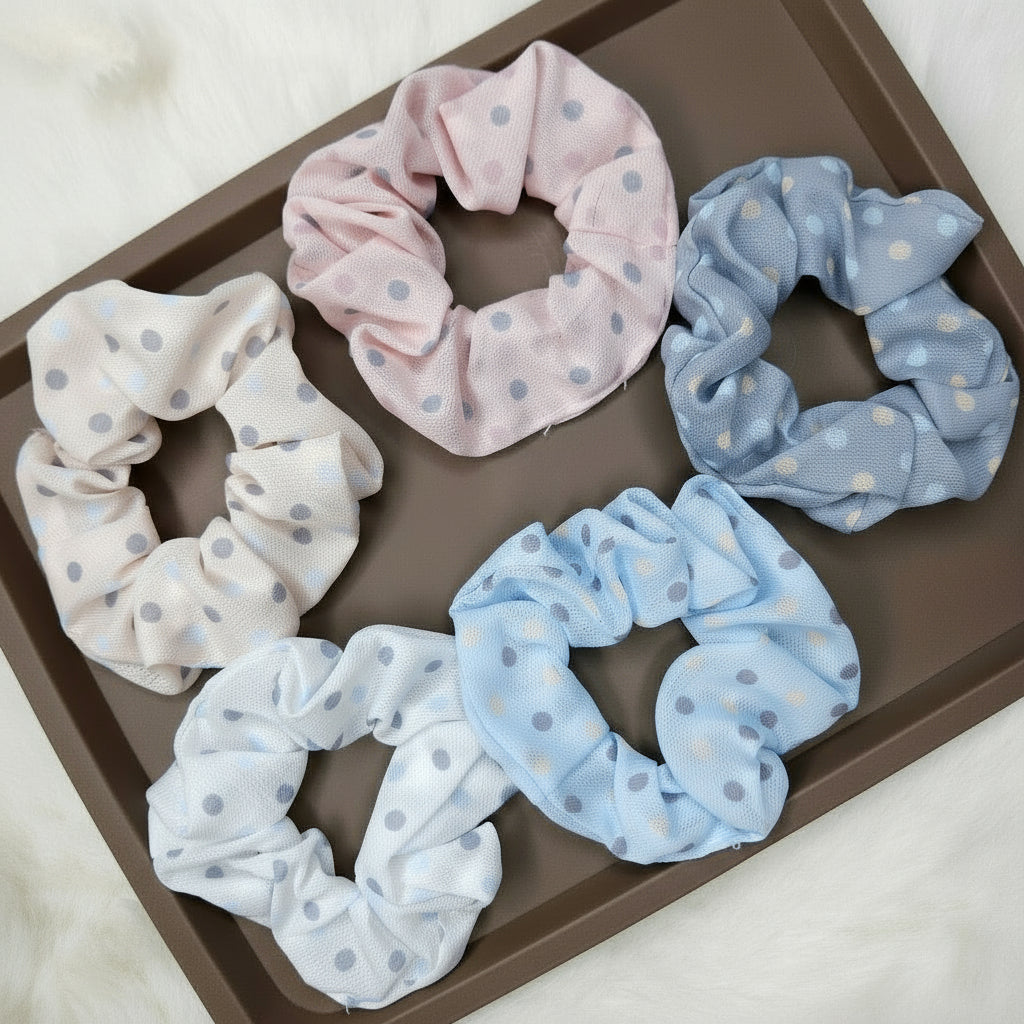 A group of five sheer mesh fabric scrunchies featuring a small polka-dot pattern. The colors are soft pastel tones: dusty pink, light blue, dusty blue-gray, cream, and white, displayed on a rectangular brown tray.