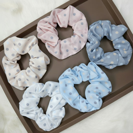 A group of five sheer mesh fabric scrunchies featuring a small polka-dot pattern. The colors are soft pastel tones: dusty pink, light blue, dusty blue-gray, cream, and white, displayed on a rectangular brown tray.