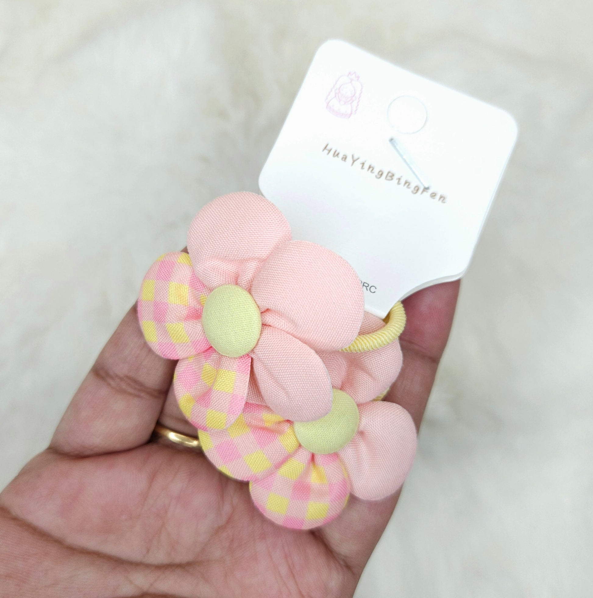 Pink and yellow flower-shaped hair tie held by a hand with a brand label in the background.