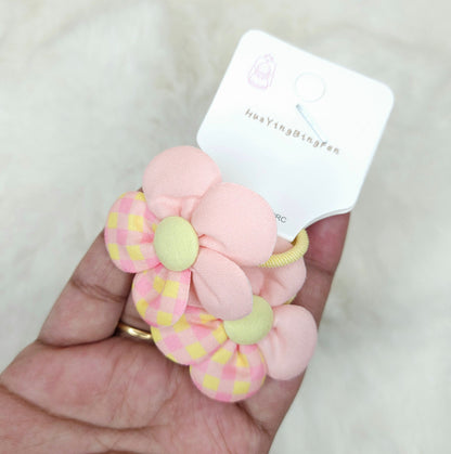 Pink and yellow flower-shaped hair tie held by a hand with a brand label in the background.