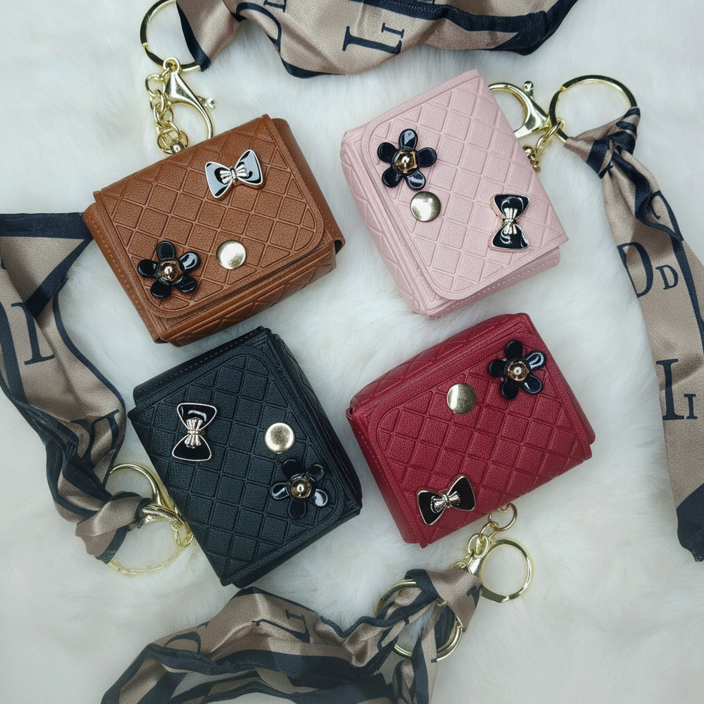 A set of four small, quilted faux leather mini-wallets used as keychains, resting on a white faux fur surface. The wallets come in brown, pink, black, and red. Each is decorated with a floral charm and a black enamel bow, and is attached to a gold-tone keyring and a long, narrow satin ribbon/scarf with a repeating pattern.