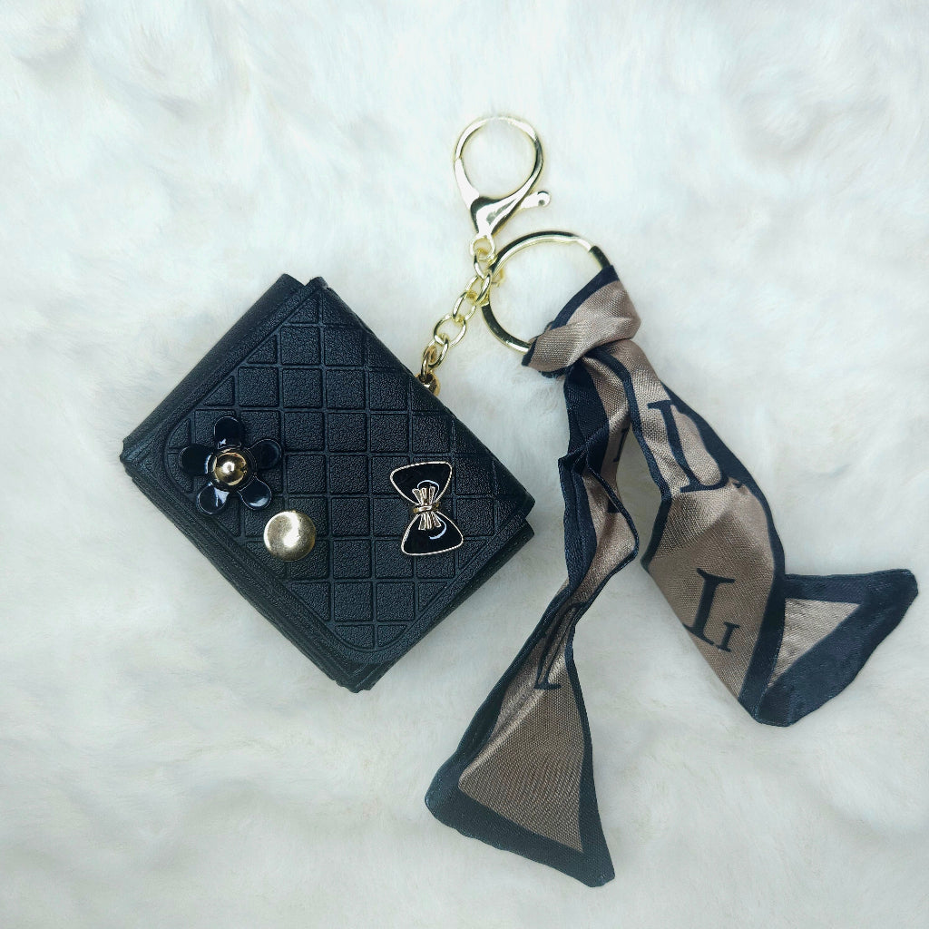 Close-up of a single Black Faux Leather Quilted Keychain Wallet. It is accessorized with a gold-tone keyring, decorative bow and flower charms, and a silky patterned scarf tie.