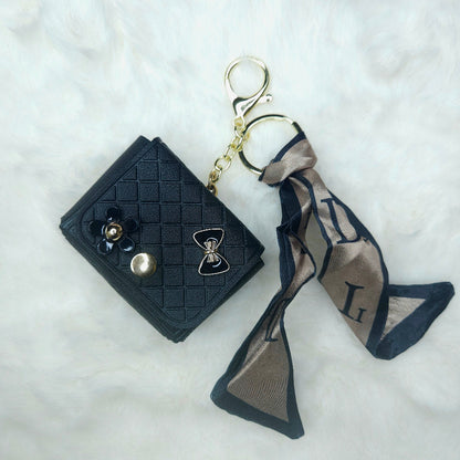 Close-up of a single Black Faux Leather Quilted Keychain Wallet. It is accessorized with a gold-tone keyring, decorative bow and flower charms, and a silky patterned scarf tie.
