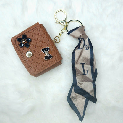A Brown Quilted Faux Leather Mini Key Pouch keychain resting on a white faux fur background. The small wallet features a diamond stitch pattern, is decorated with a black enamel bow and a black floral charm, and is attached to a gold-tone keyring with a long, patterned satin scarf ribbon.