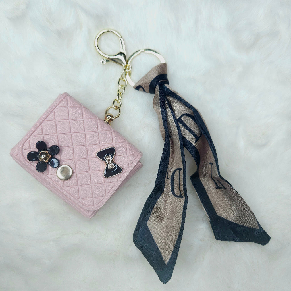 Small Light Pink Faux Leather Key Wallet with gold-tone metal clasp and ring. Features a decorative black bow and flower charm. Includes a silky scarf accessory for securing to a bag or wrist.