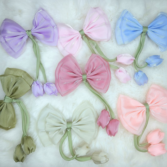 A flat lay image of six sheer fabric bows in pastel colors (lavender, light pink, light blue, medium pink, olive green, and cream) arranged on a white fluffy surface. Each bow has a long green stem-like attachment with two small, closed fabric tulip buds dangling from it in a matching pastel shade.